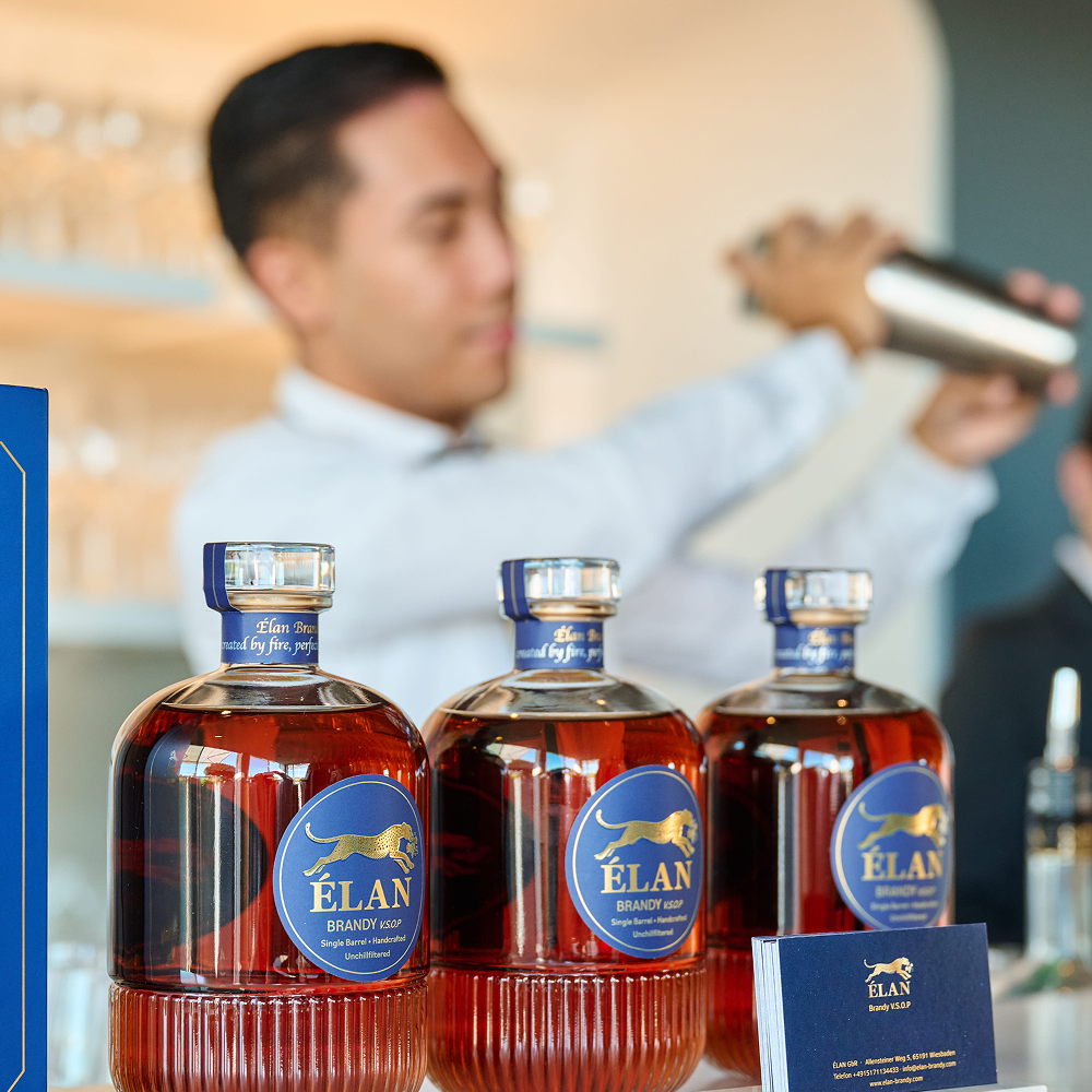Drei Flaschen Elan VSOP Brandy, im Hintergrund ein Barkeeper, der einen Cocktail mixt.