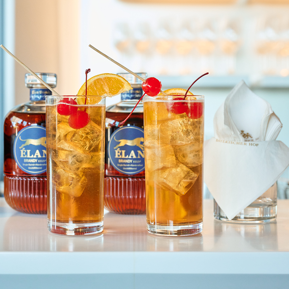 Zwei eisgekühlte Cocktails, garniert mit Orangenscheiben und Maraschino-Kirschen, vor zwei Flaschen Élan-Brandy.