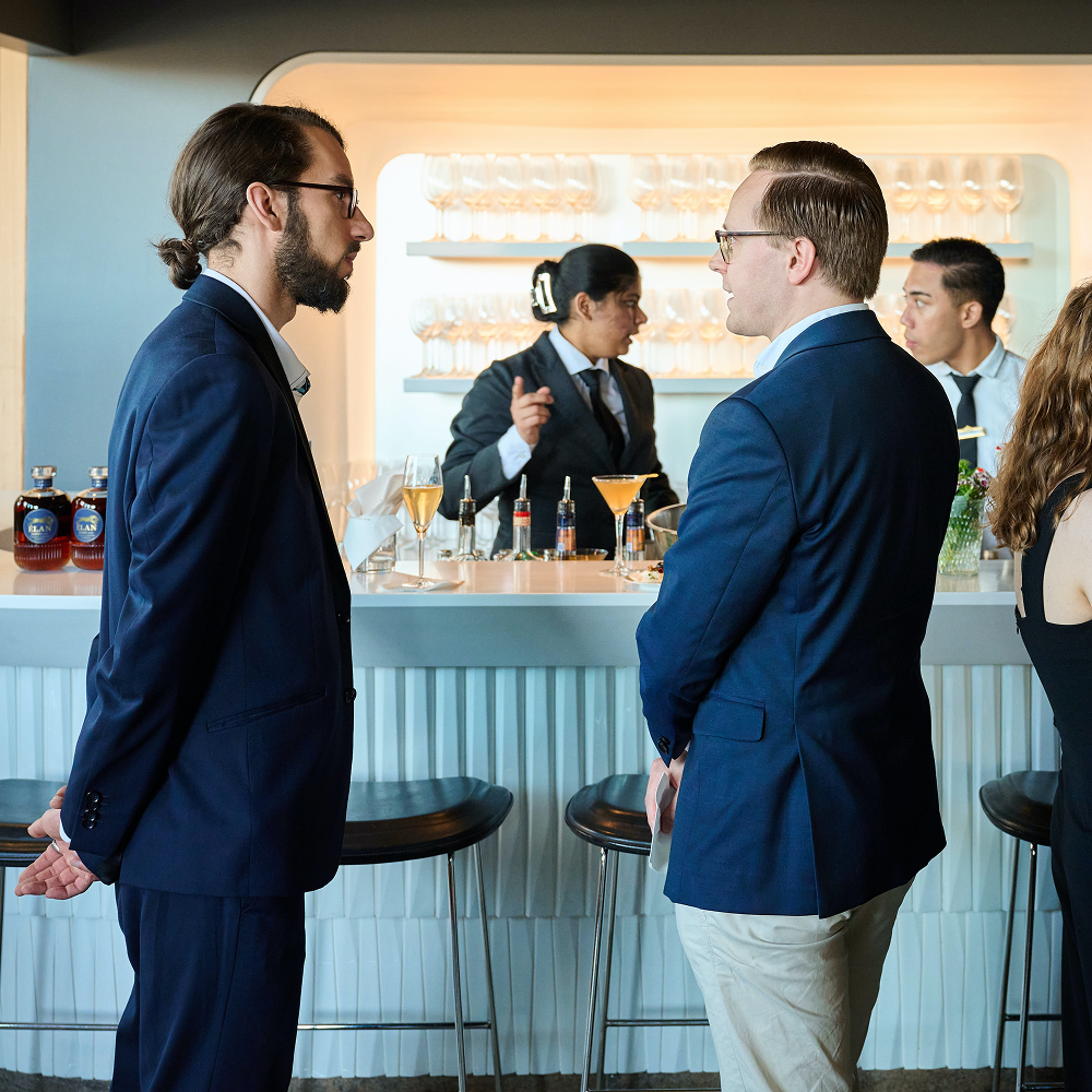 Zwei Männer in Anzügen unterhalten sich an einer Bar, während ein Kellner hinter der Theke steht.