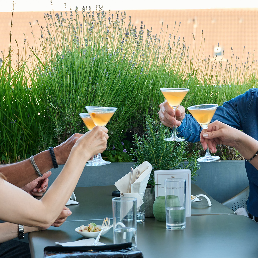 Drei Personen heben an einem Tisch im Freien ihre Martini-Gläser, um anzustoßen.