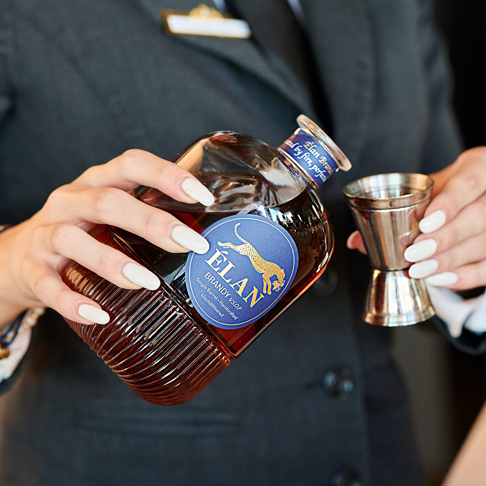Ein Mann im Anzug gießt Élan Brandy VSOP aus einer Glasflasche in einen silbernen Jigger.