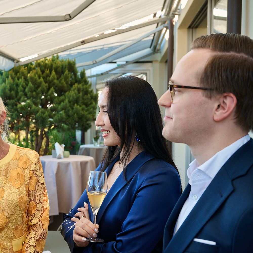 Drei Personen bei einer Veranstaltung im Freien; in der Mitte steht eine Frau, die ein Glas Champagner in der Hand hält.